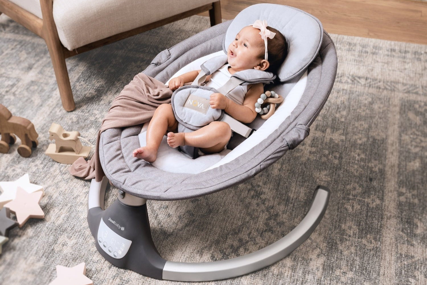 Baby in a gray baby swing on a carpeted floor with toys around
