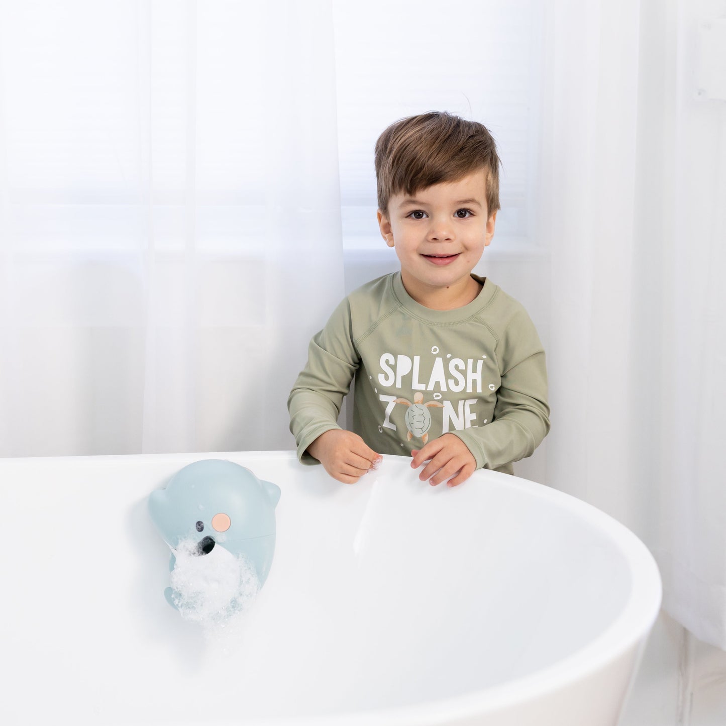 Child sitting on a white bathtub with a toy, wearing a shirt with text, against a white curtain background.