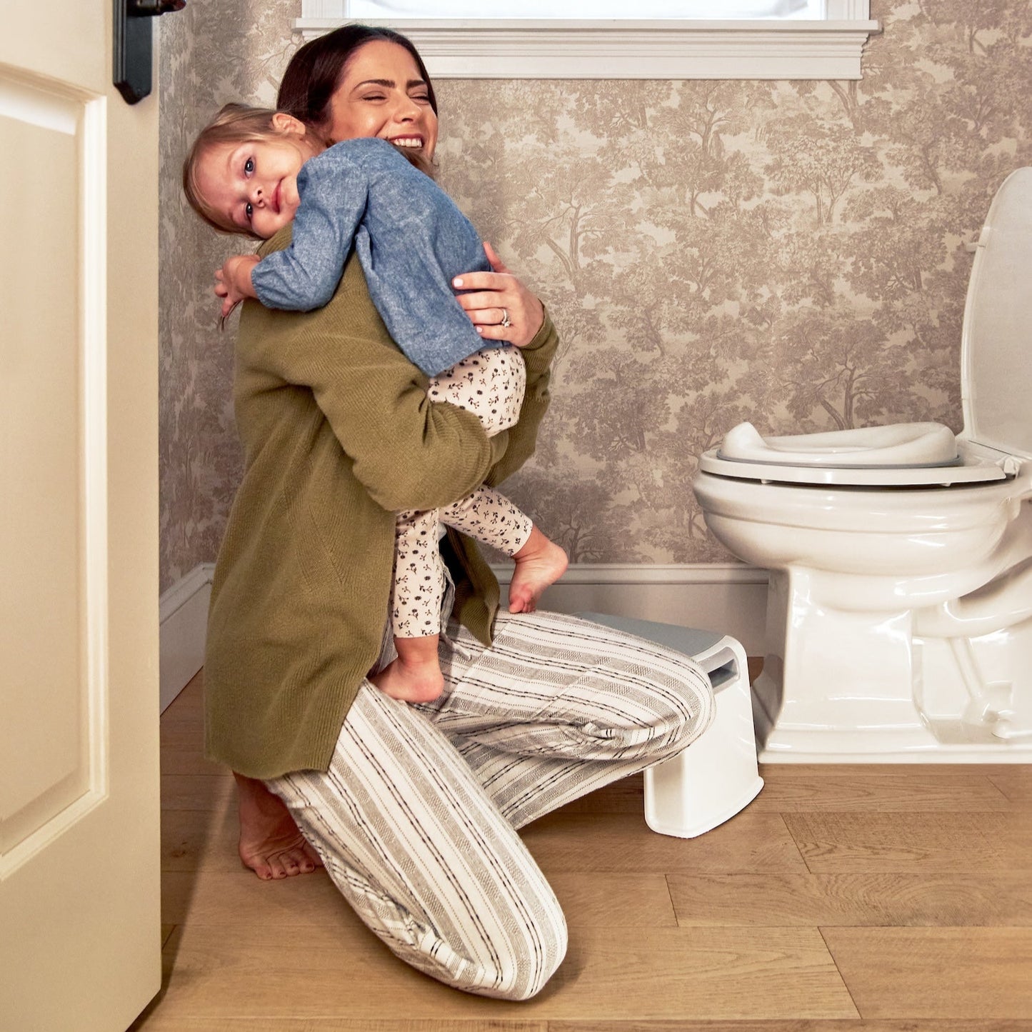 Woman holding a baby in a bathroom with a toilet and window in the background.