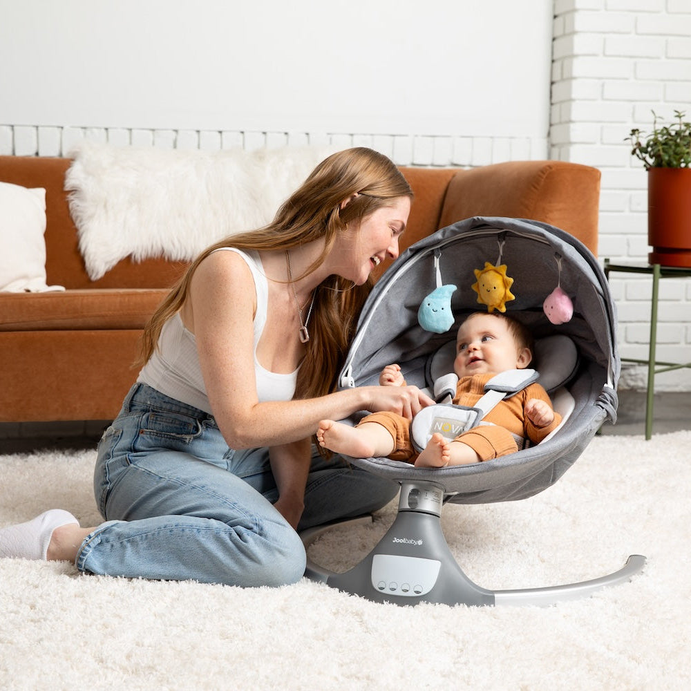 Woman sitting on the floor with a baby in a baby swing in a living room.
