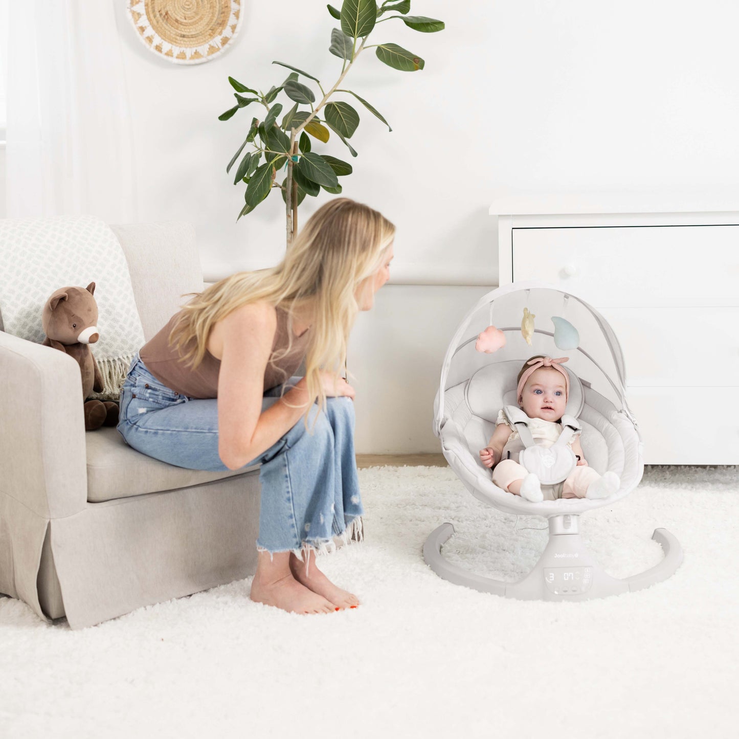 Woman sitting on a couch next to a baby in a swing chair in a bright room.