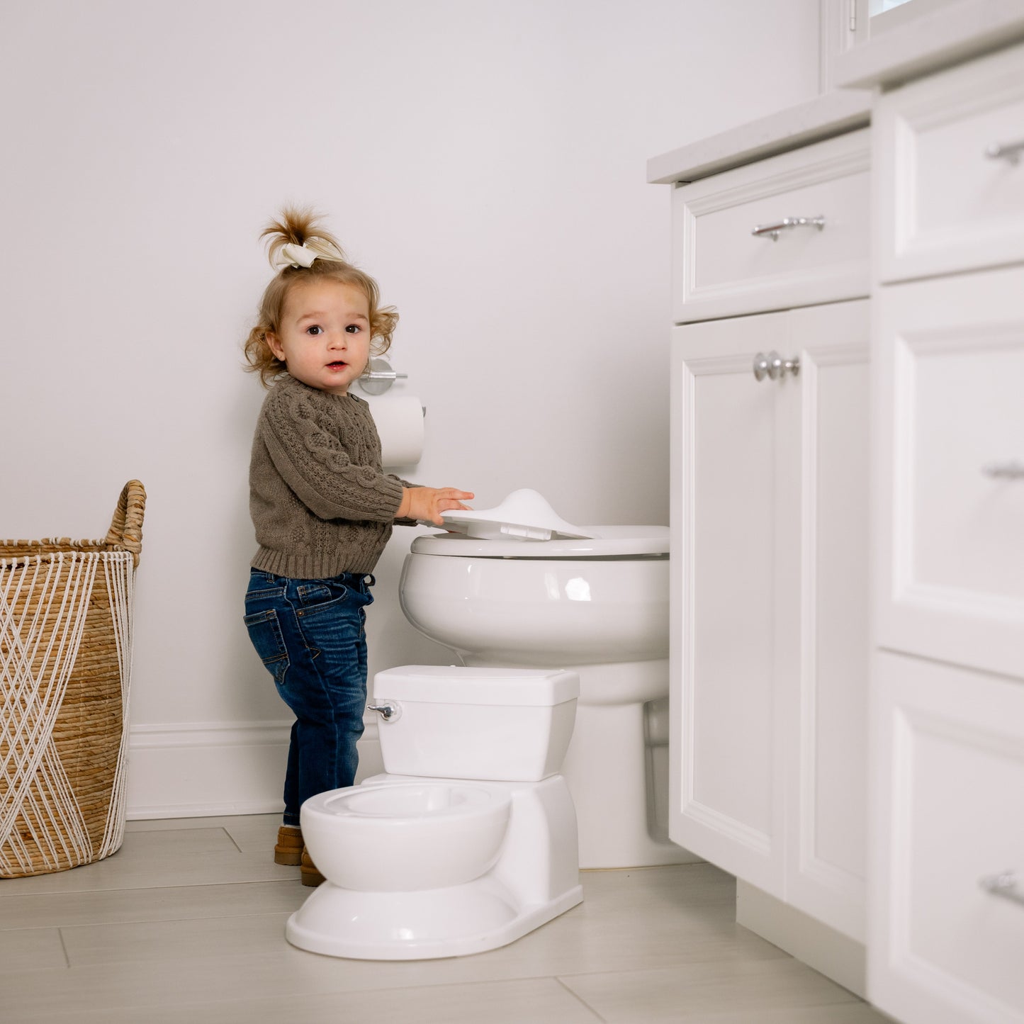 Child standing next to a toilet in a bathroom with white cabinets and a wicker basket.