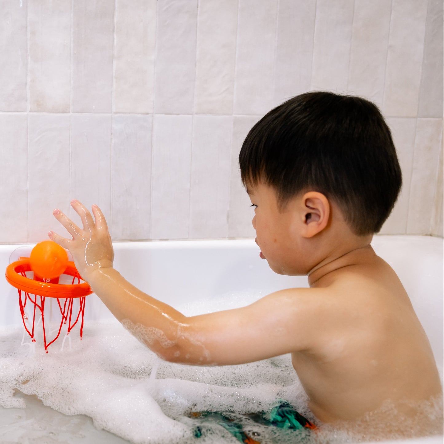 Child playing with a basketball hoop in a bathtub filled with water and bubbles.