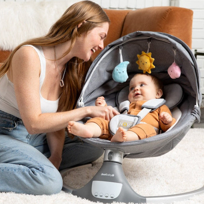 Woman sitting on the floor with a baby in a baby swing in a living room.