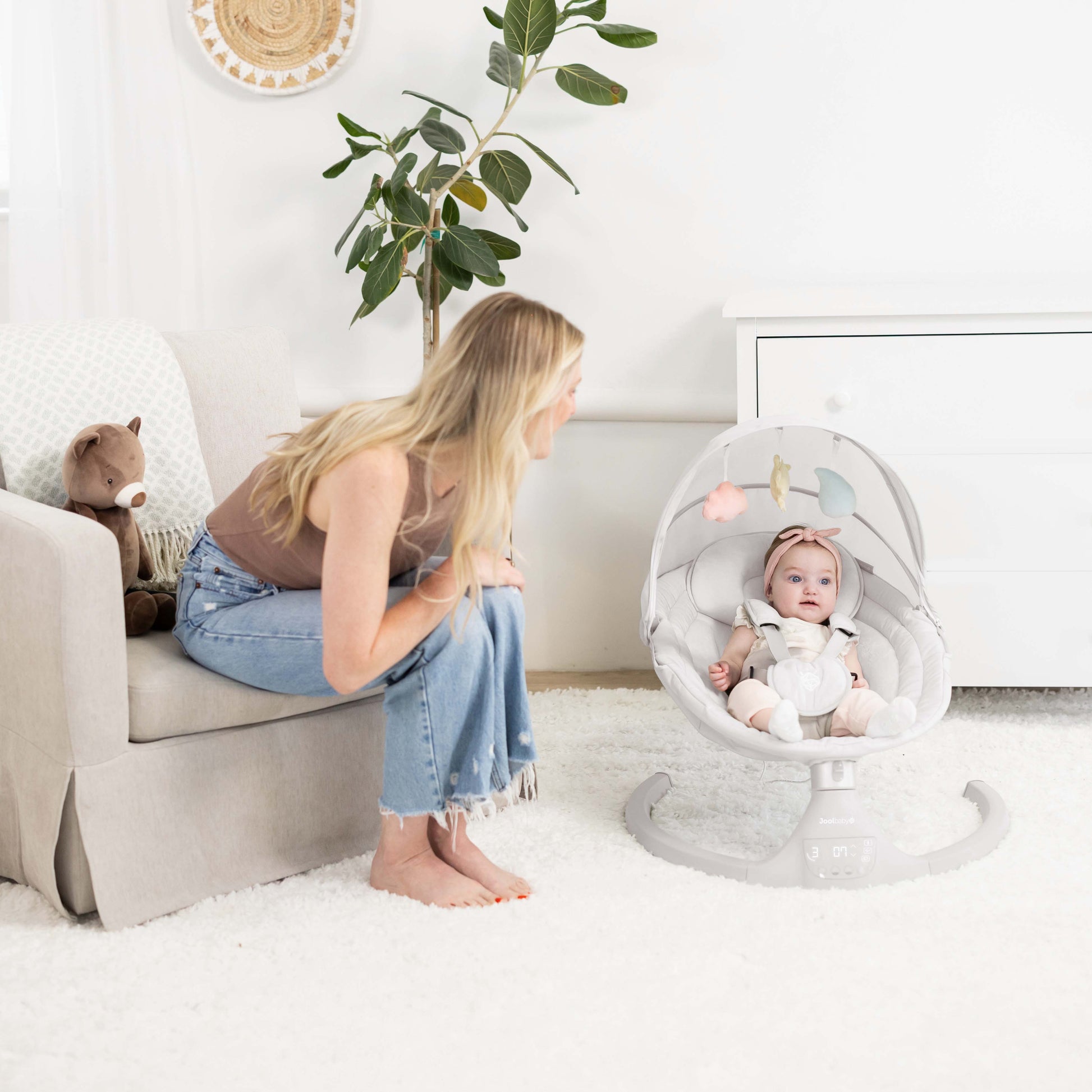 Woman sitting on a couch next to a baby in a swing chair in a bright room.