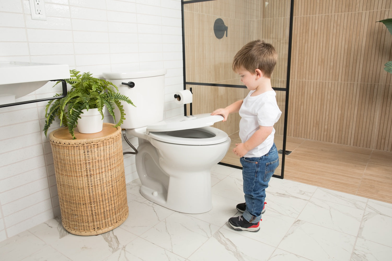 Child using a toilet with a wicker stool and plant in a bathroom.
