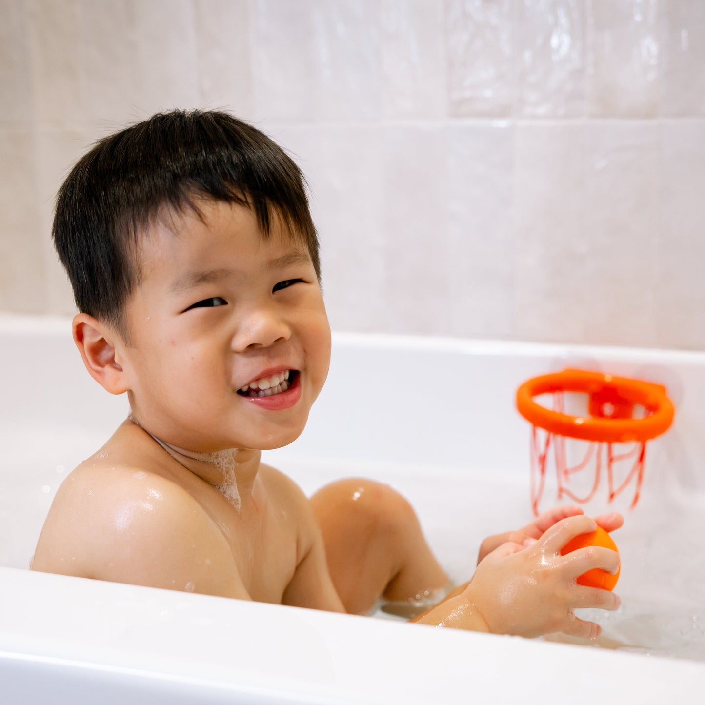 Child playing with a basketball toy in a bathtub, smiling.